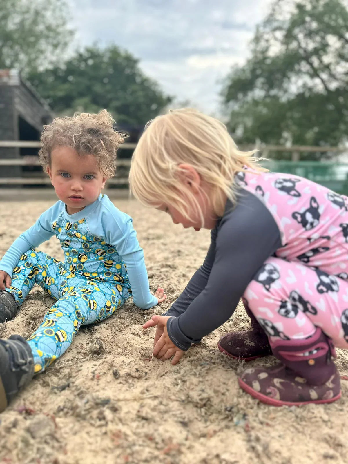Two young children wearing colourful riding base layer sets - one in a blue bee print and the other in a pink dog print - playing in the sand at a riding arena.