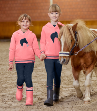Two young children in matching coral riding hoodies holding hands and leading a small pony inside an arena.
